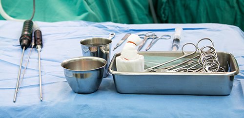 Surgical tools including forceps, clamps, a scalpel, and a stainless steel bowl on a sterile blue medical drape.